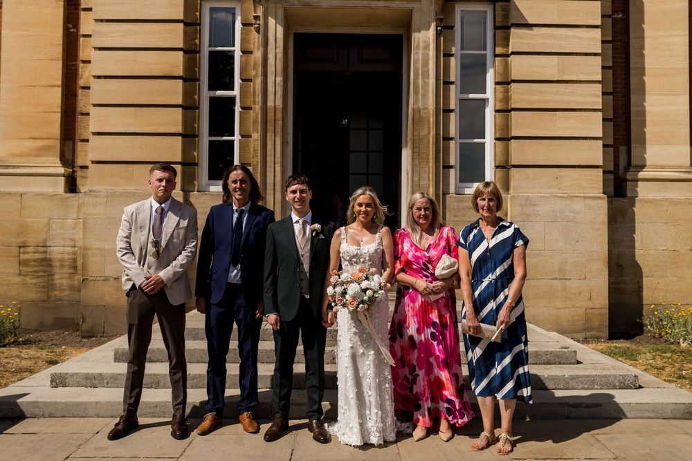 Wedding party group photo outside grand stone building on sunny day with bride holding bouquet.
