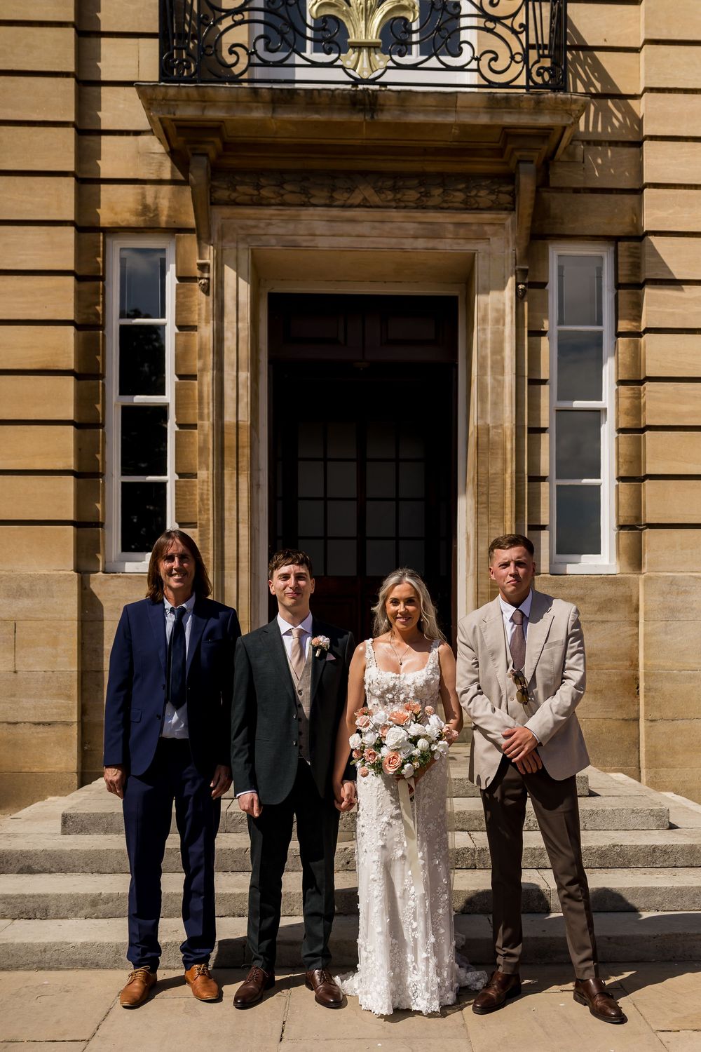 Bride and groom pose with two groomsmen outside elegant stone building entrance on bright sunny day.
