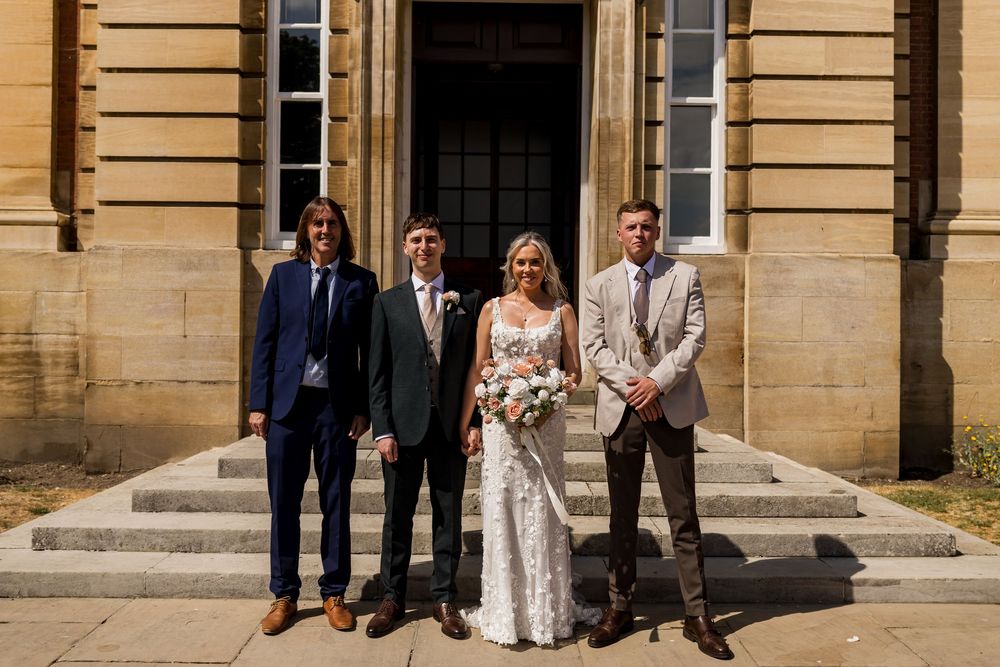 Bride with bouquet posing with three groomsmen outside grand stone building wedding venue.