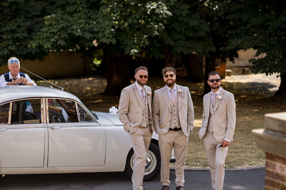 Three groomsmen in beige suits posing beside vintage white wedding car on sunny day.