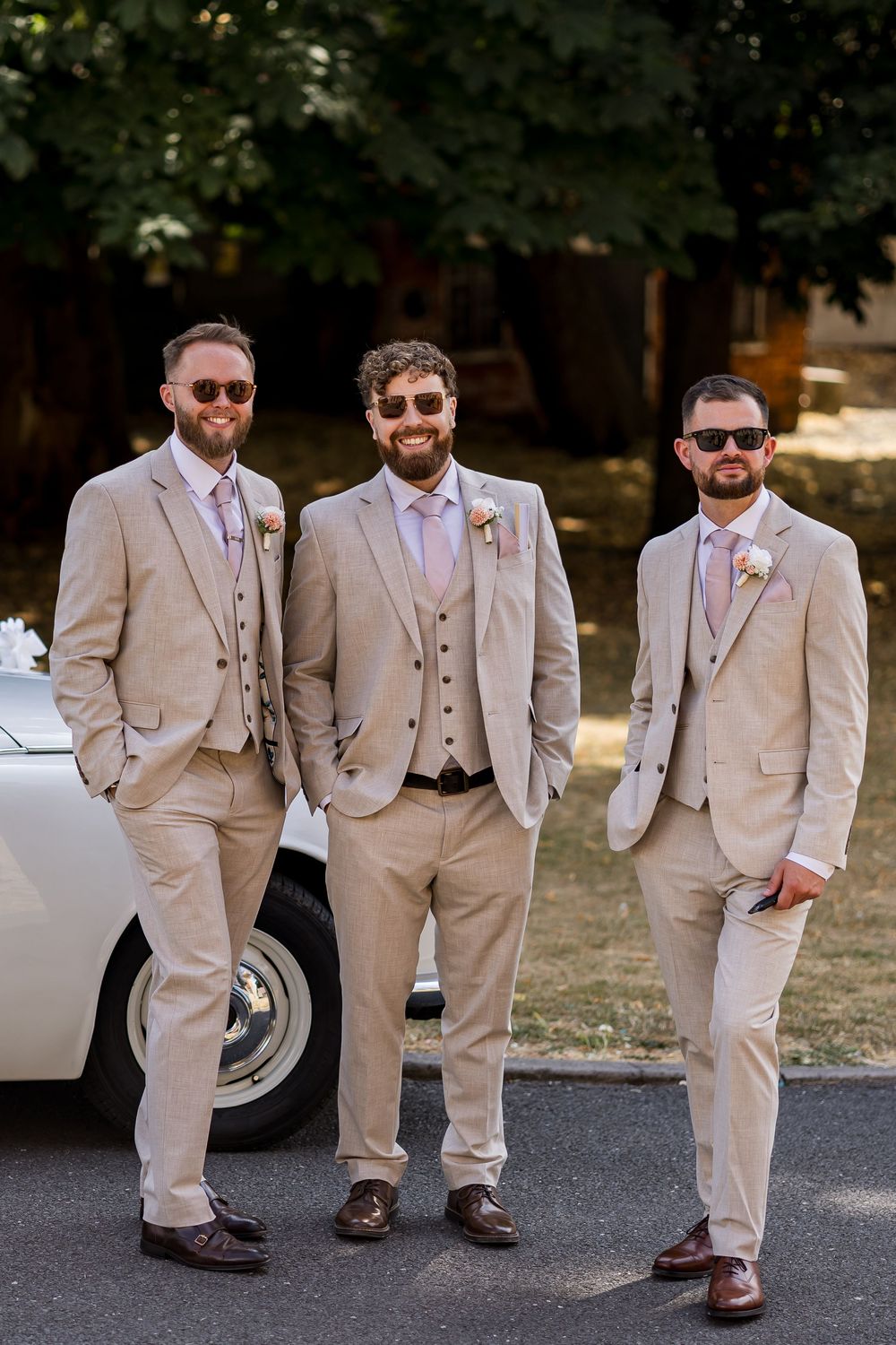 Three groomsmen in matching tan suits posing beside vintage white car outdoors.