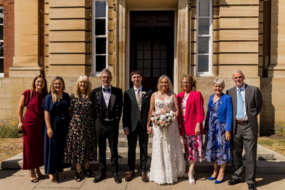 Wedding couple pose with family members outside a stone building on a sunny wedding day.