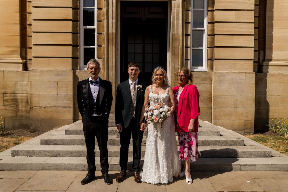 Bride and groom pose with two family members on steps of a stunning classical stone building on wedding day.