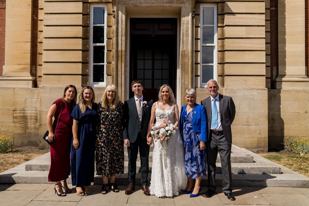 Bride and groom celebrate with six family members outside a beautiful classical stone building on their wedding day.
