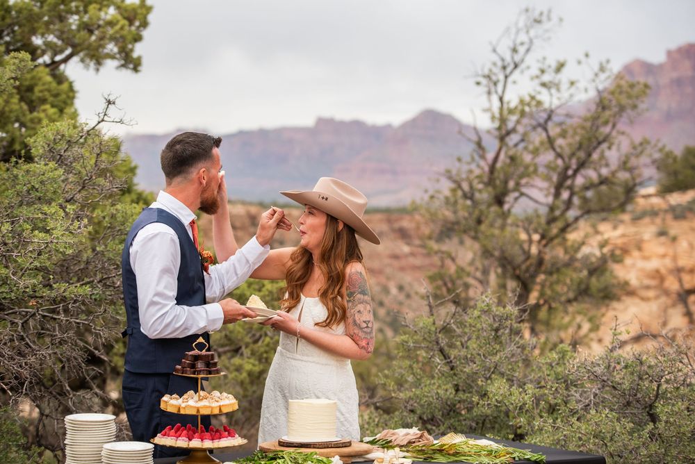 Couple sharing cake outdoors at desert wedding with red rock canyon views and rustic table spread.