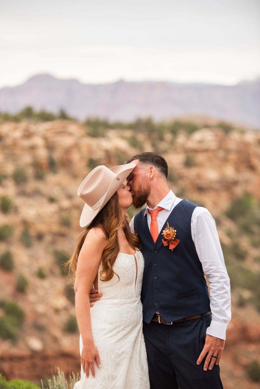Bride and groom share a kiss at a desert overlook, she wears a wide-brim hat, he wears a navy vest with orange boutonniere.