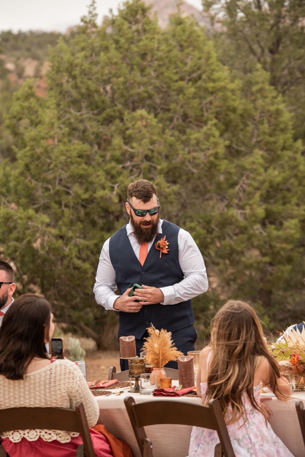 Bearded groom in navy vest and orange tie giving a speech at an outdoor wedding reception.