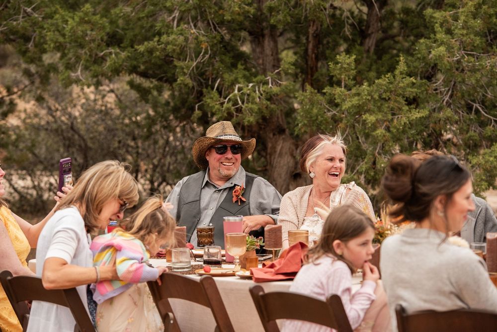 Guests laughing and enjoying an outdoor wedding reception dinner at rustic wooden tables surrounded by trees.