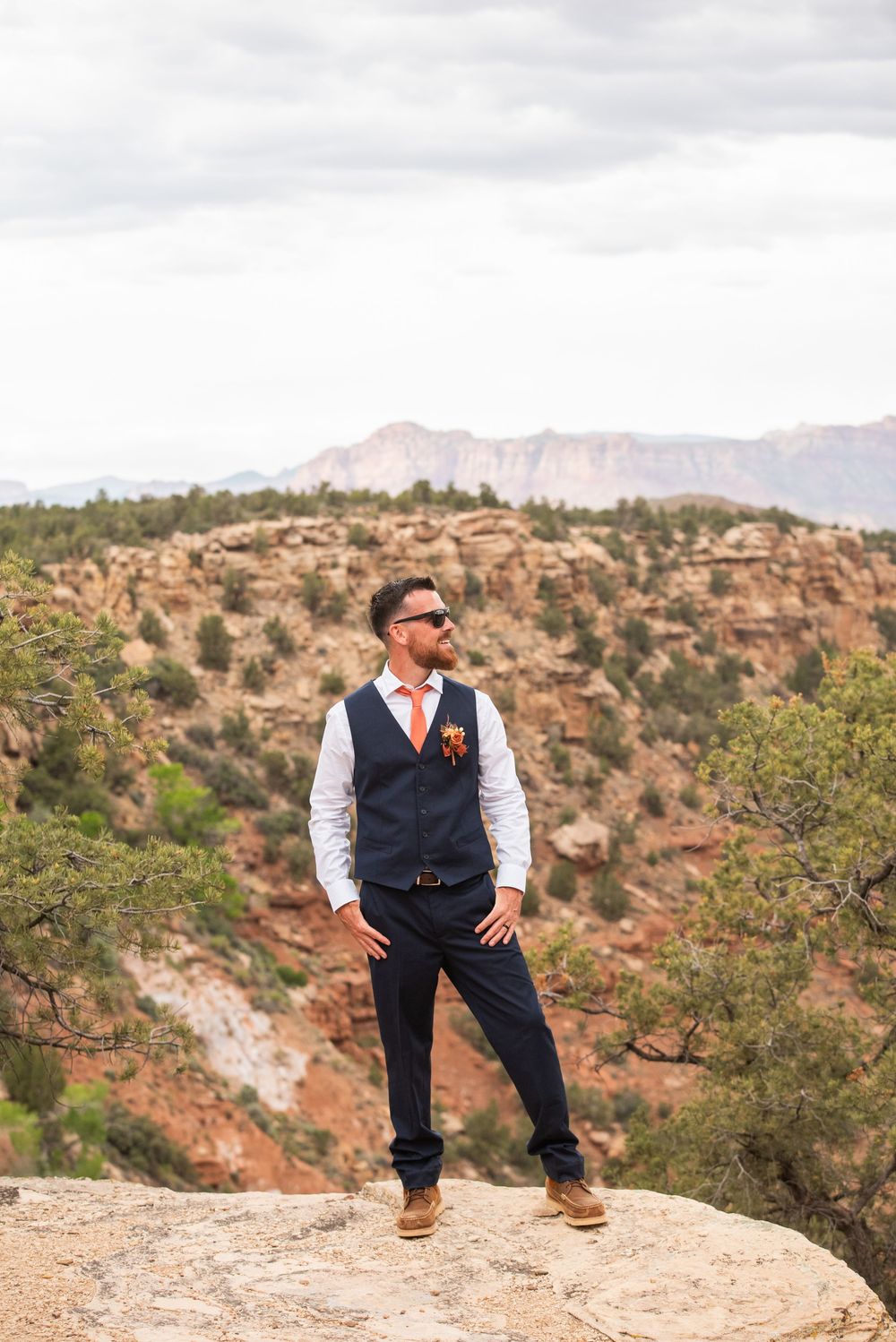Groom in navy vest and orange tie poses on rocky cliff with red canyon landscape behind him.