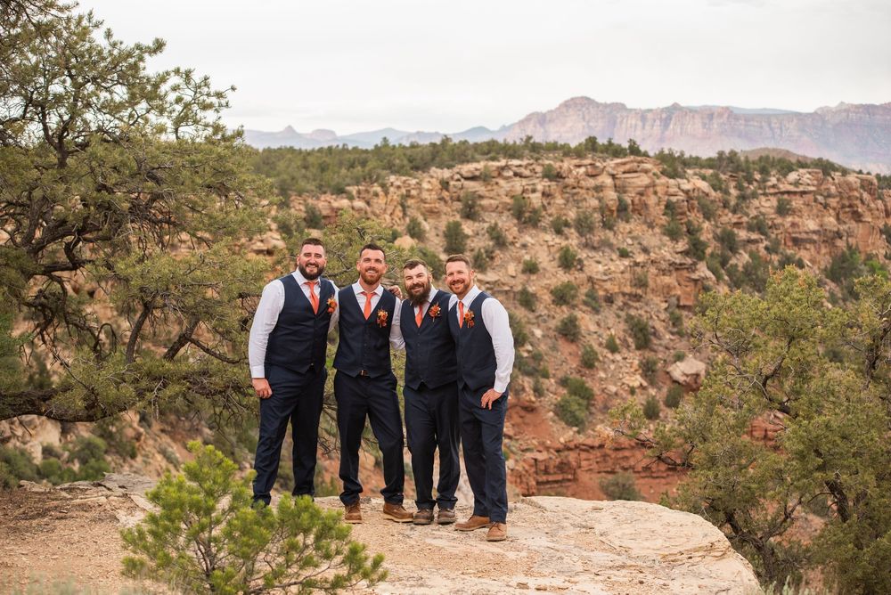 Four groomsmen in navy vests and orange ties pose on a rocky overlook with red canyon mountains in the background.