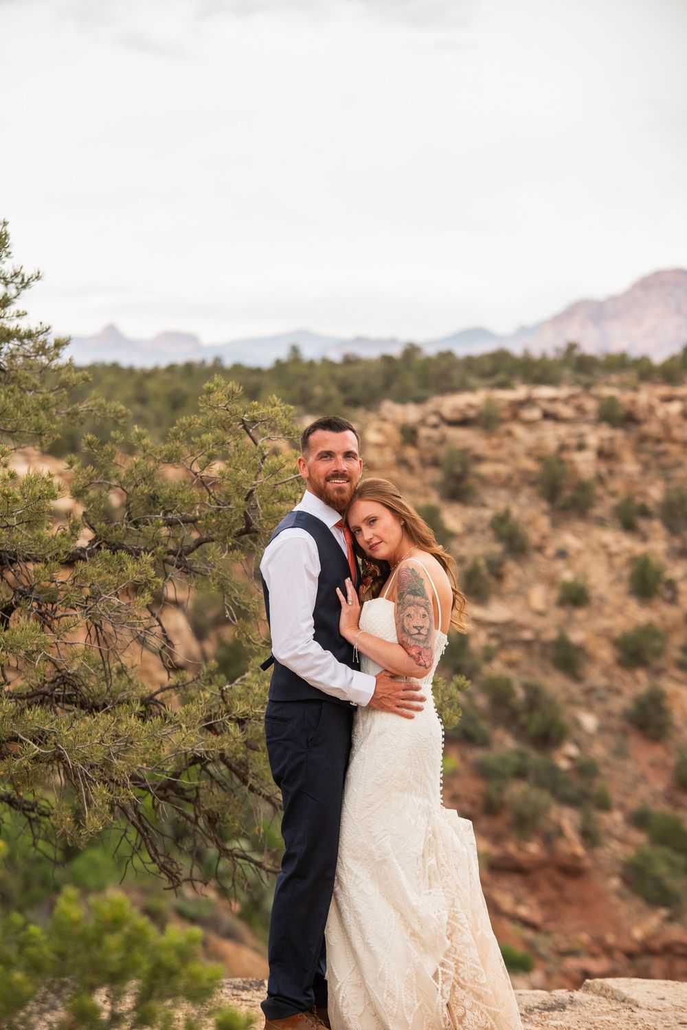Bride and groom embrace on a rocky overlook with red canyon landscape and evergreen trees in the background.