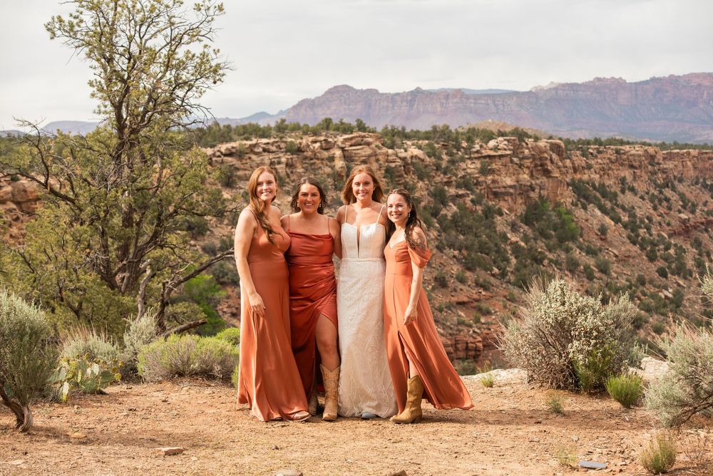 Bride and three bridesmaids in rust-orange dresses pose together outdoors with red rock canyon landscape backdrop.