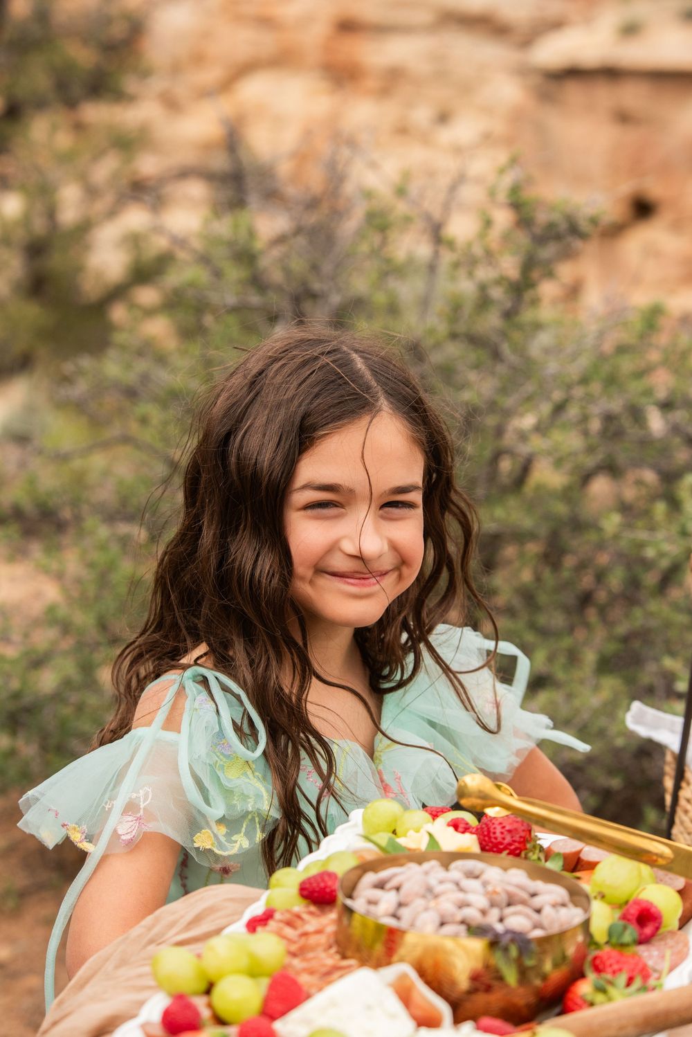 Young girl in mint dress smiling at outdoor picnic spread with fresh fruits and snacks in a rocky landscape.