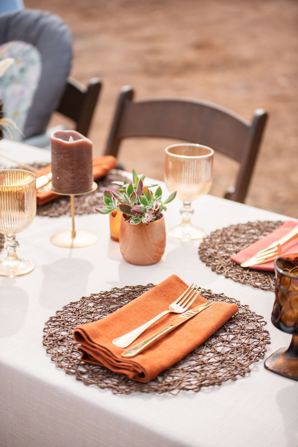 Elegant fall wedding table setting with burnt orange napkins, gold cutlery, woven placemats, and candles on a white tablecloth.
