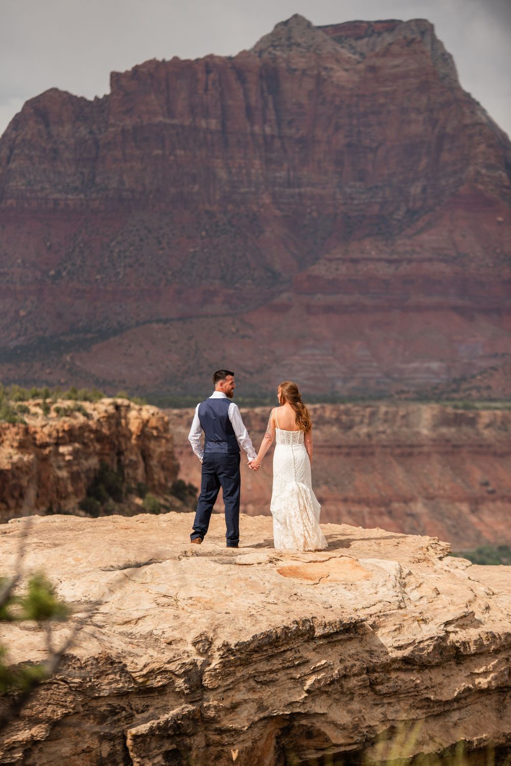Bride and groom hold hands on rocky cliff overlooking dramatic red rock canyon landscape.