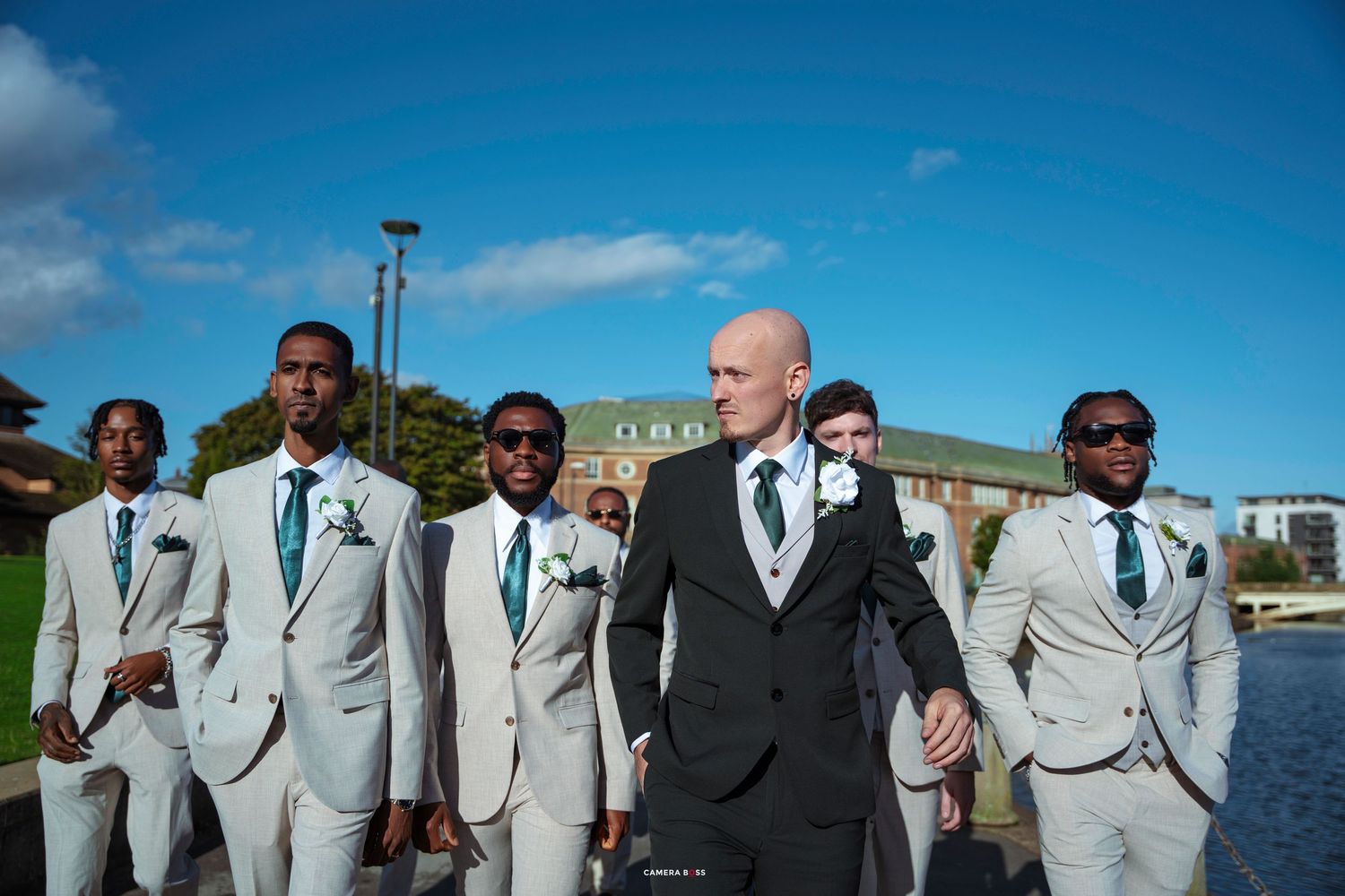 Groomsmen in light gray suits with teal ties walk outdoors near a pond on a sunny wedding day.