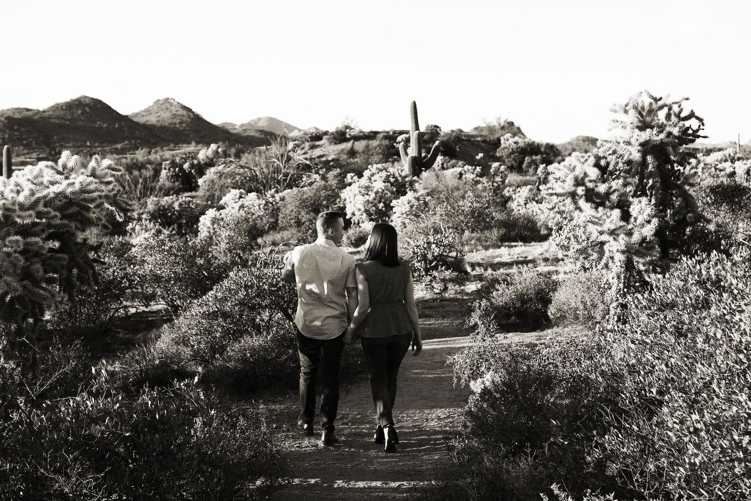 Black and white photo of a couple walking arm in arm along a desert trail lined with cacti and mountains.