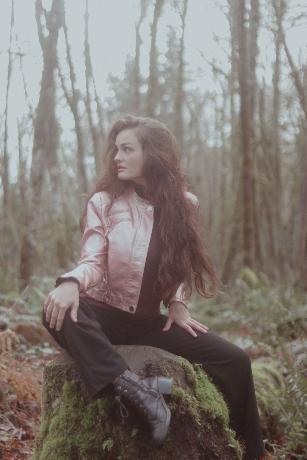 Woman with long wavy hair in a pink metallic jacket and black boots sits on a moss-covered stump in a misty forest.