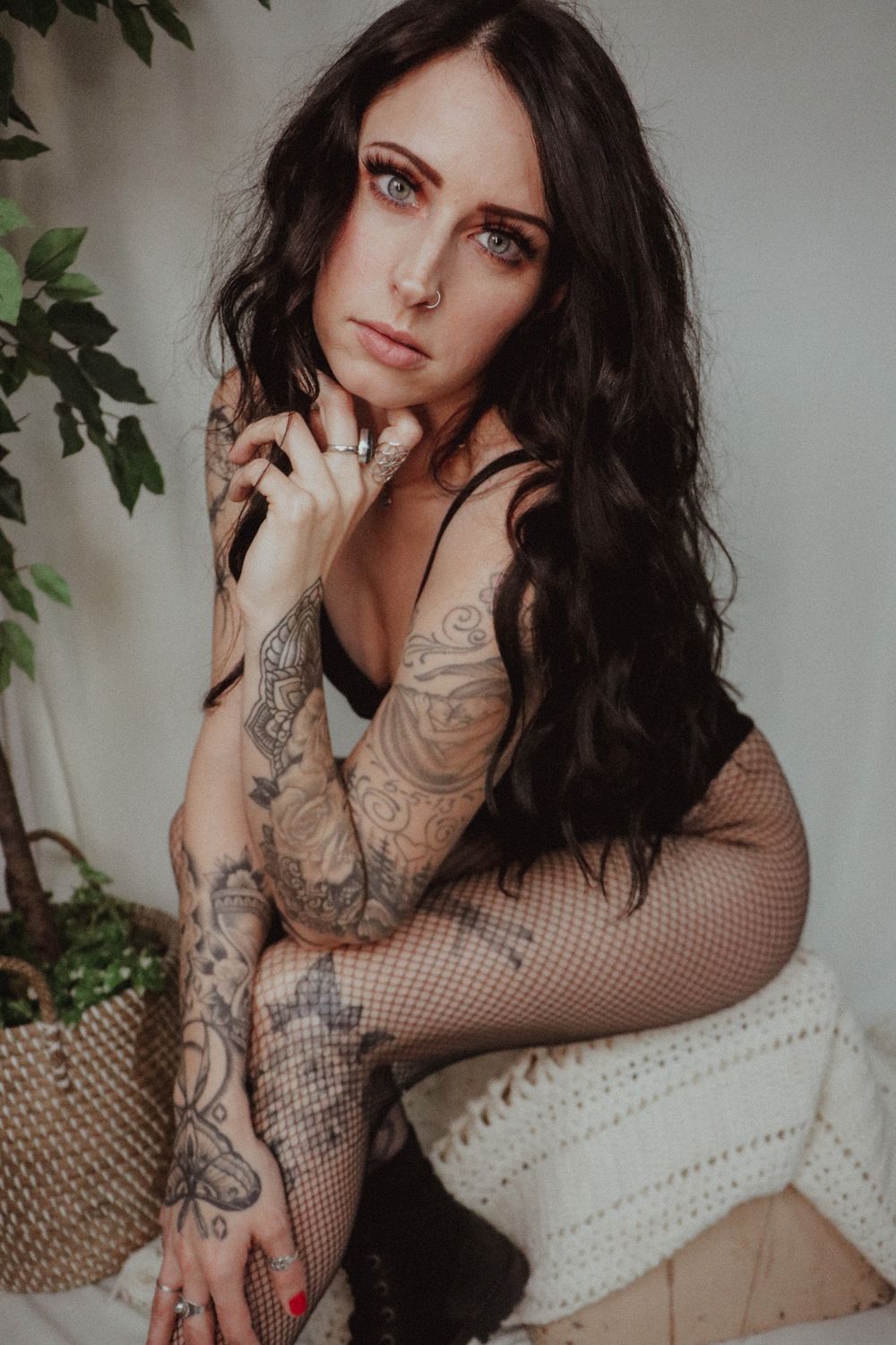Tattooed woman with long dark wavy hair and blue eyes poses seated in fishnet tights beside a potted plant in a bright studio.