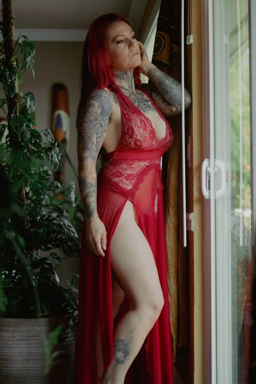 Tattooed woman with red hair in a red lace lingerie robe stands beside a window and leafy green houseplant in a boudoir portrait.