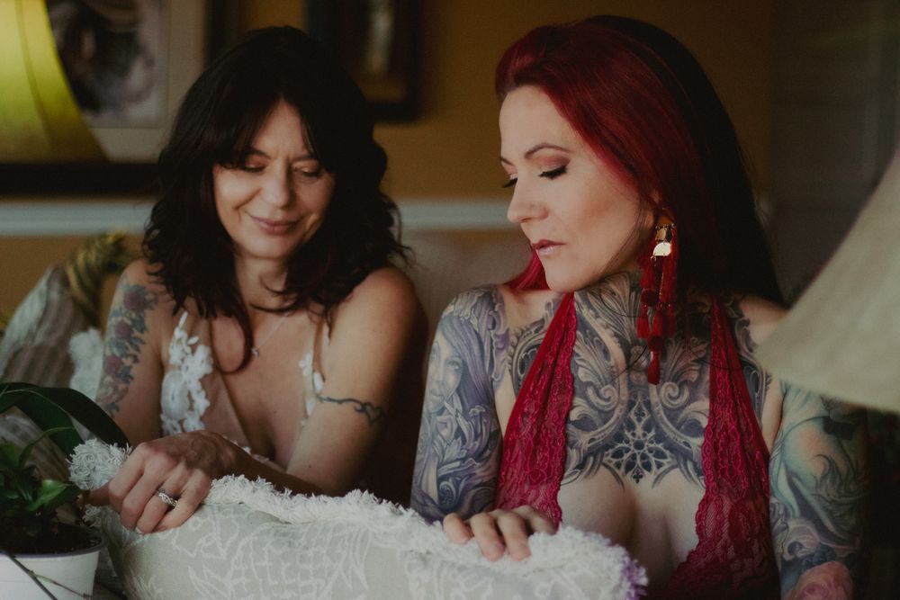 Two tattooed women sit close together on a cozy couch, one with dark wavy hair and one with red hair, gazing down at a fluffy white blanket.