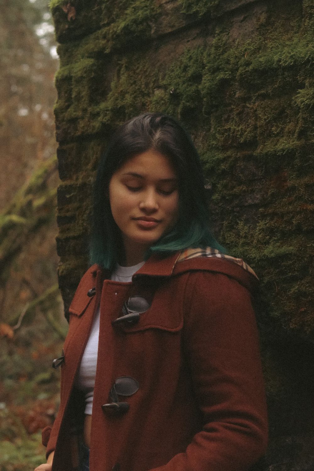 Young woman with teal-tipped dark hair and a red duffle coat stands with eyes closed beside a moss-covered tree trunk in a forest.