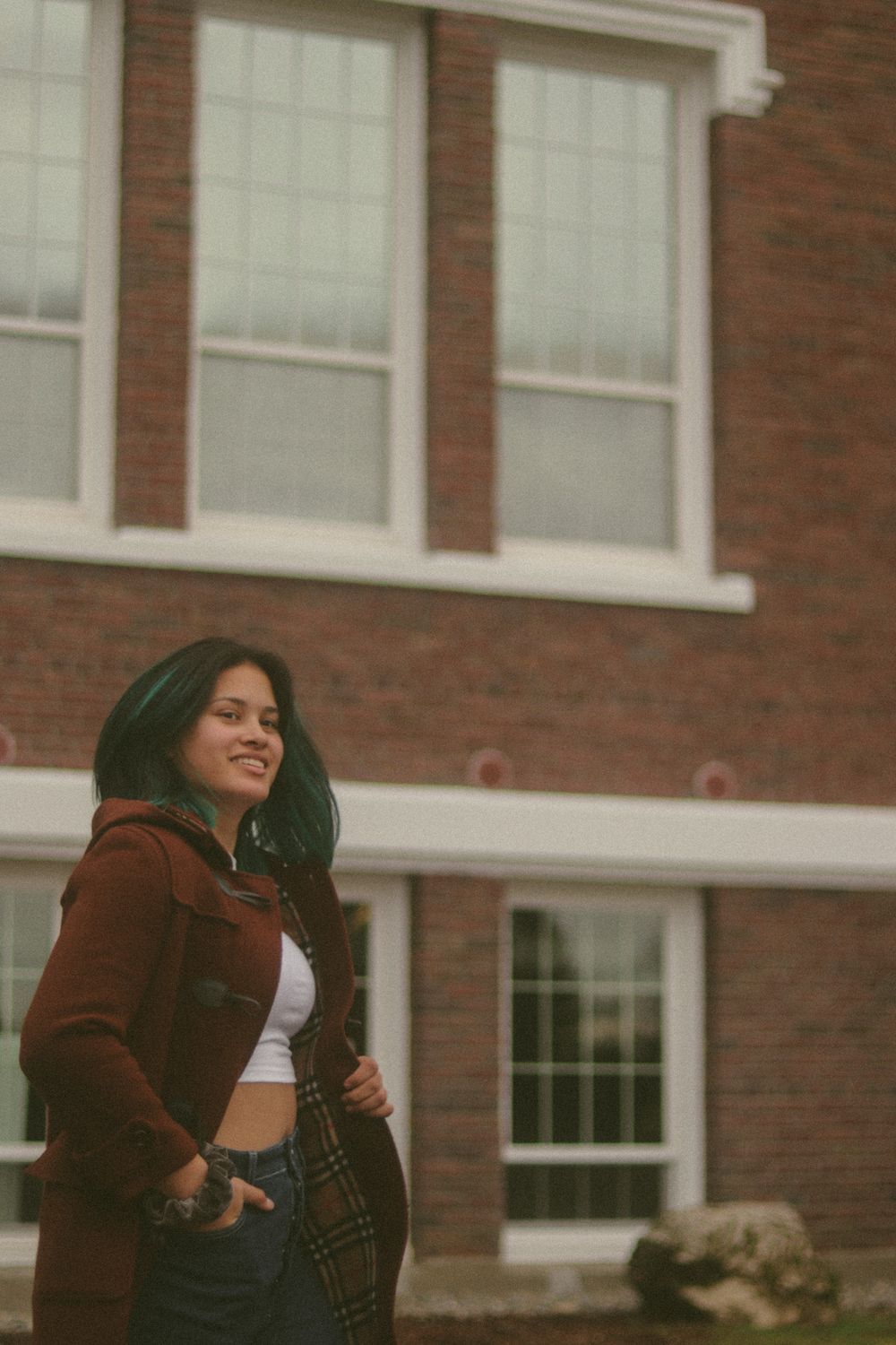 Woman with dark green-tinted hair smiles in a rust-brown coat and white crop top, standing before a red brick building with large windows.