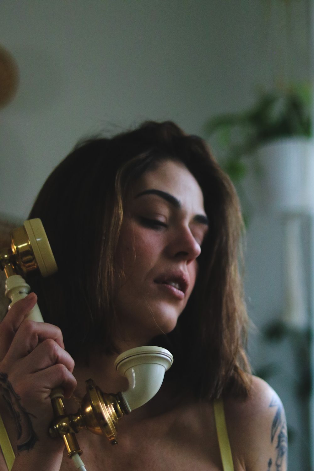 Woman with tousled brown hair and arm tattoos holds a vintage brass and cream rotary telephone receiver beside a leafy houseplant indoors.