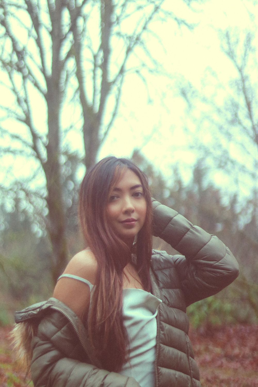 Woman with long brown hair in an olive puffer jacket and satin top poses with a hand behind her head among bare trees in soft hazy light.