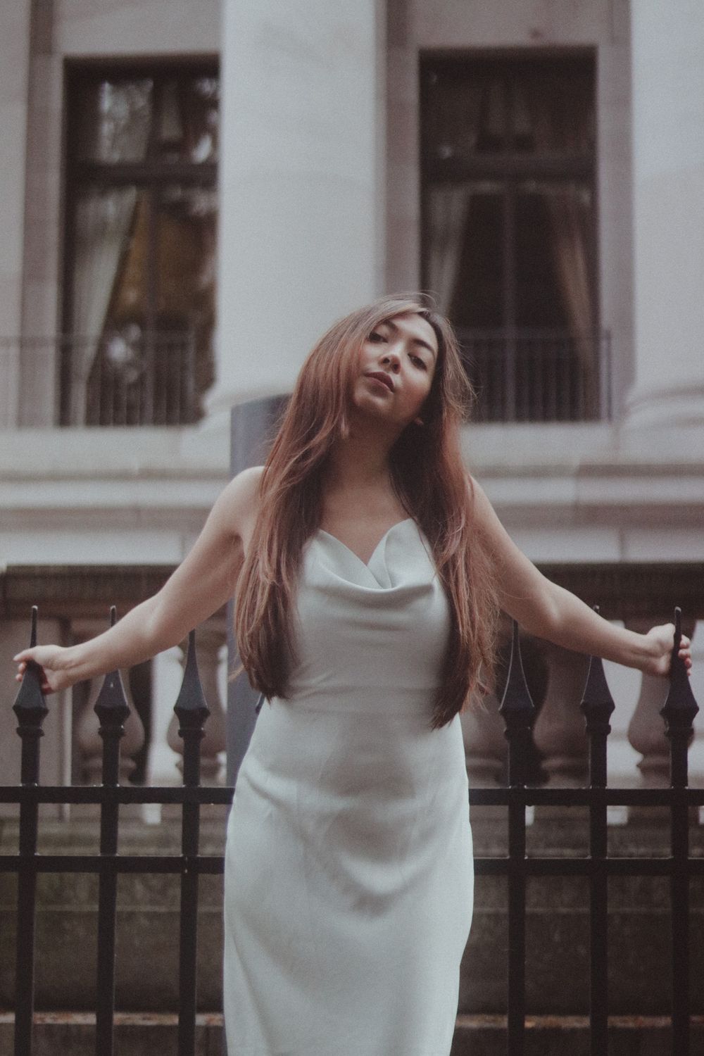 Woman with long auburn hair in a white satin dress stands with arms outstretched before an iron fence and a columned stone building.
