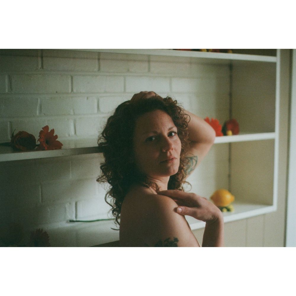 Woman with curly auburn hair looks over her bare shoulder beside white shelves holding flowers and fruit in soft natural light.