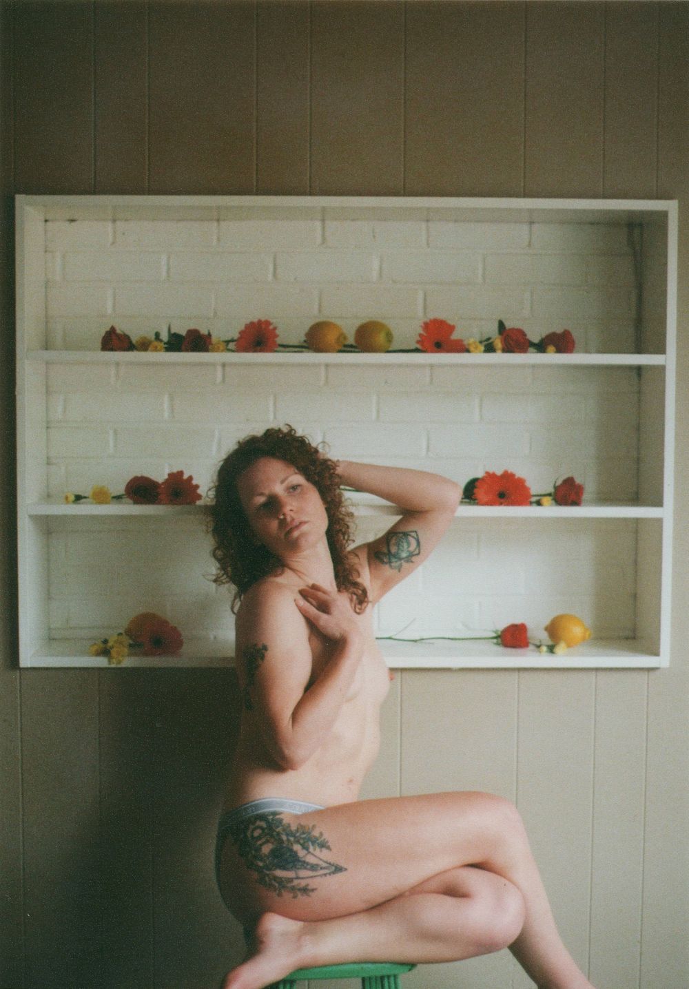 Woman with curly hair and tattoos sits on a green stool in front of white shelves decorated with orange flowers and fruit.
