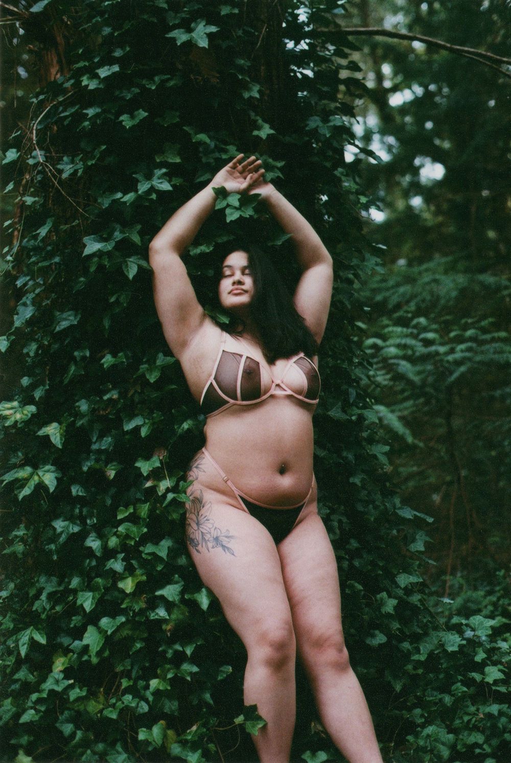 Woman in a mesh bralette and briefs stands with arms raised before a tree covered in green ivy in a forest.