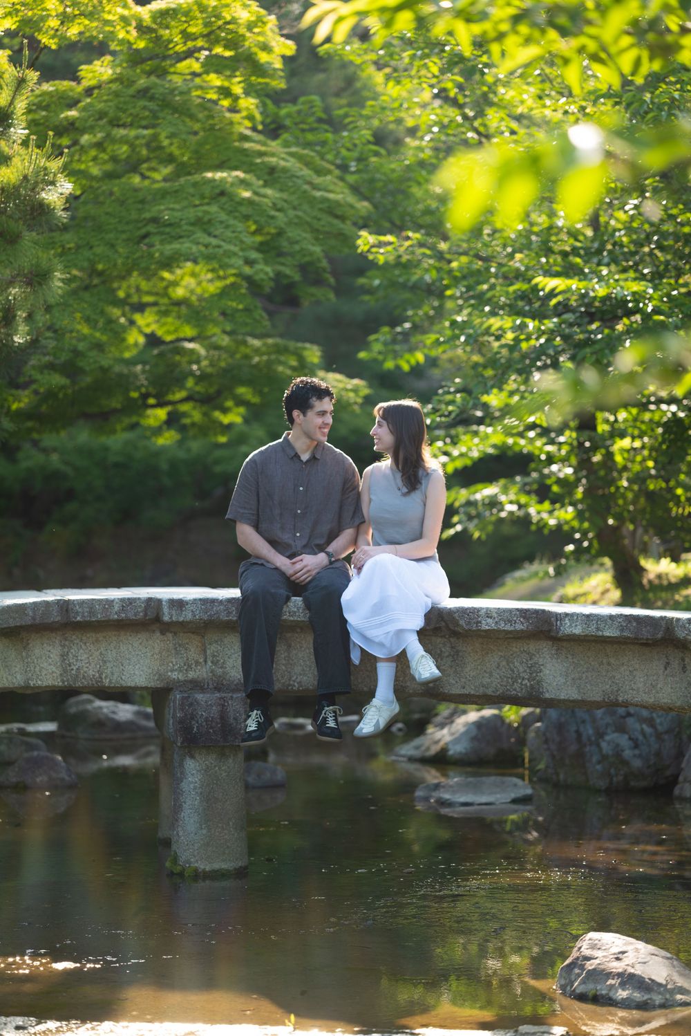 A couple sits together on a stone bridge over a tranquil stream, surrounded by lush green trees in a serene park.