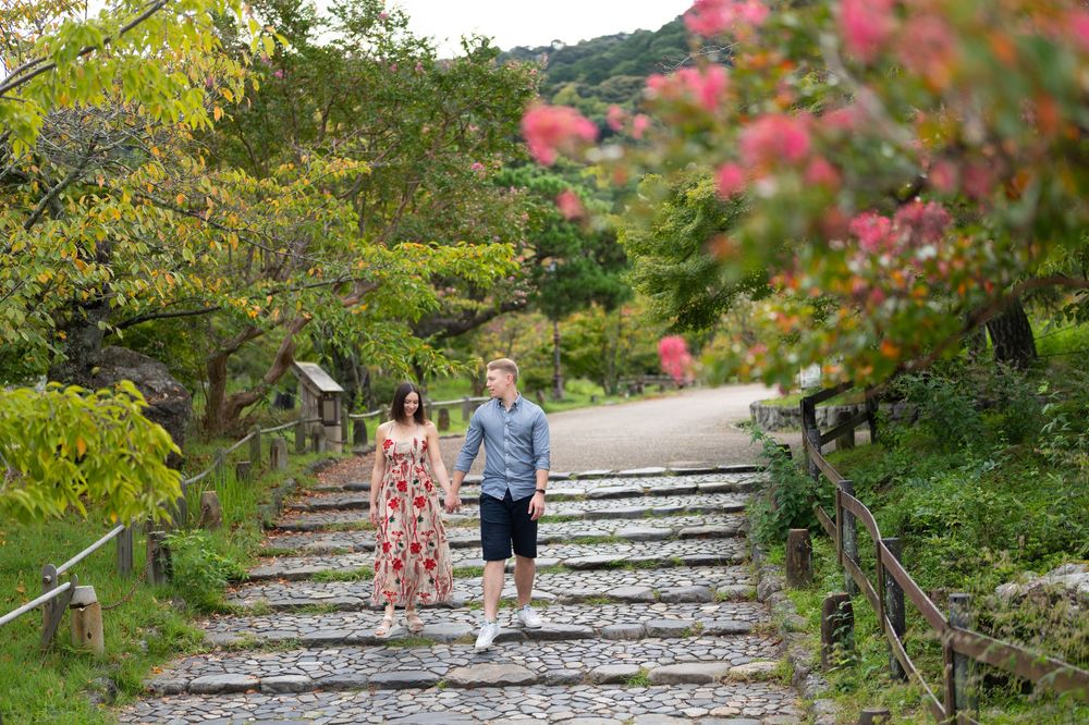 Couple walking hand-in-hand down stone steps surrounded by lush greenery and pink flowering bushes in a Japanese garden.