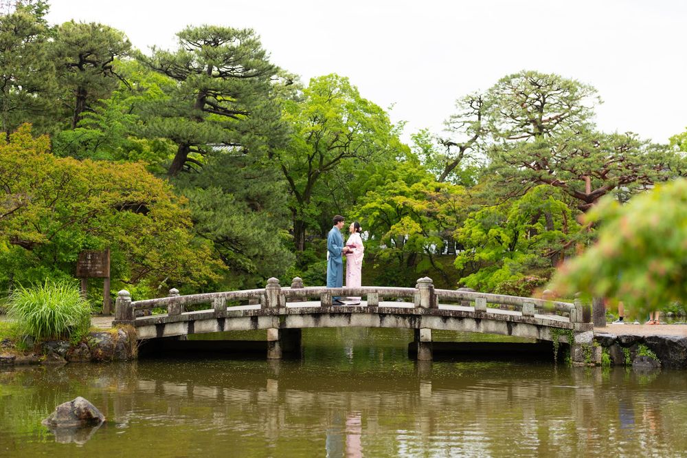 Couple in traditional kimono standing on a stone arched bridge in a lush Japanese garden with a tranquil pond.