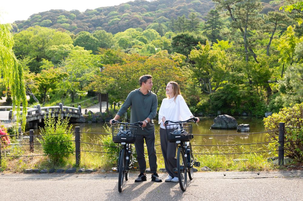 Couple standing with bicycles on a scenic park path surrounded by lush green trees and a tranquil pond.