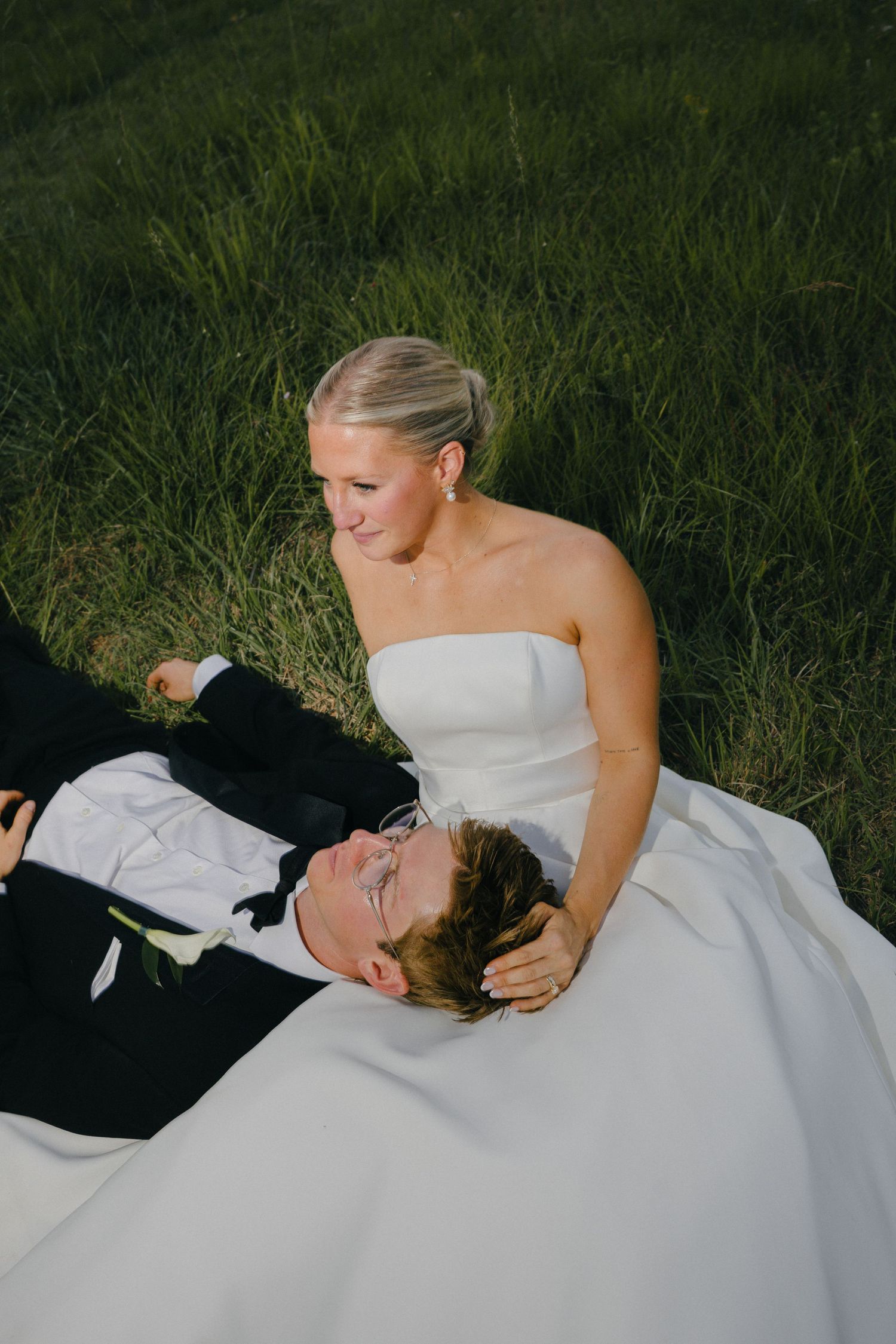 Bride in strapless white gown sits on grass while groom in black tuxedo lays in her lap during outdoor wedding photos.