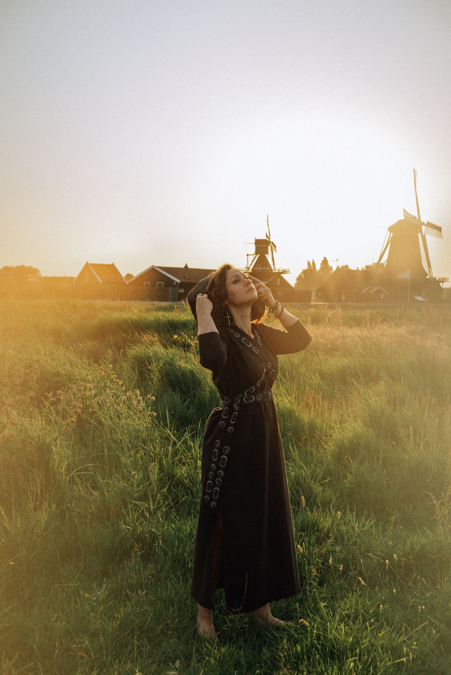 Woman in a long black dress stands barefoot in a green grassy field at golden sunset with traditional Dutch windmills behind her.