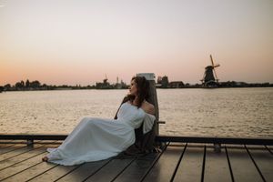 Woman in a flowing white dress sits on a wooden dock at sunset, overlooking calm water and a traditional Dutch windmill in the distance.