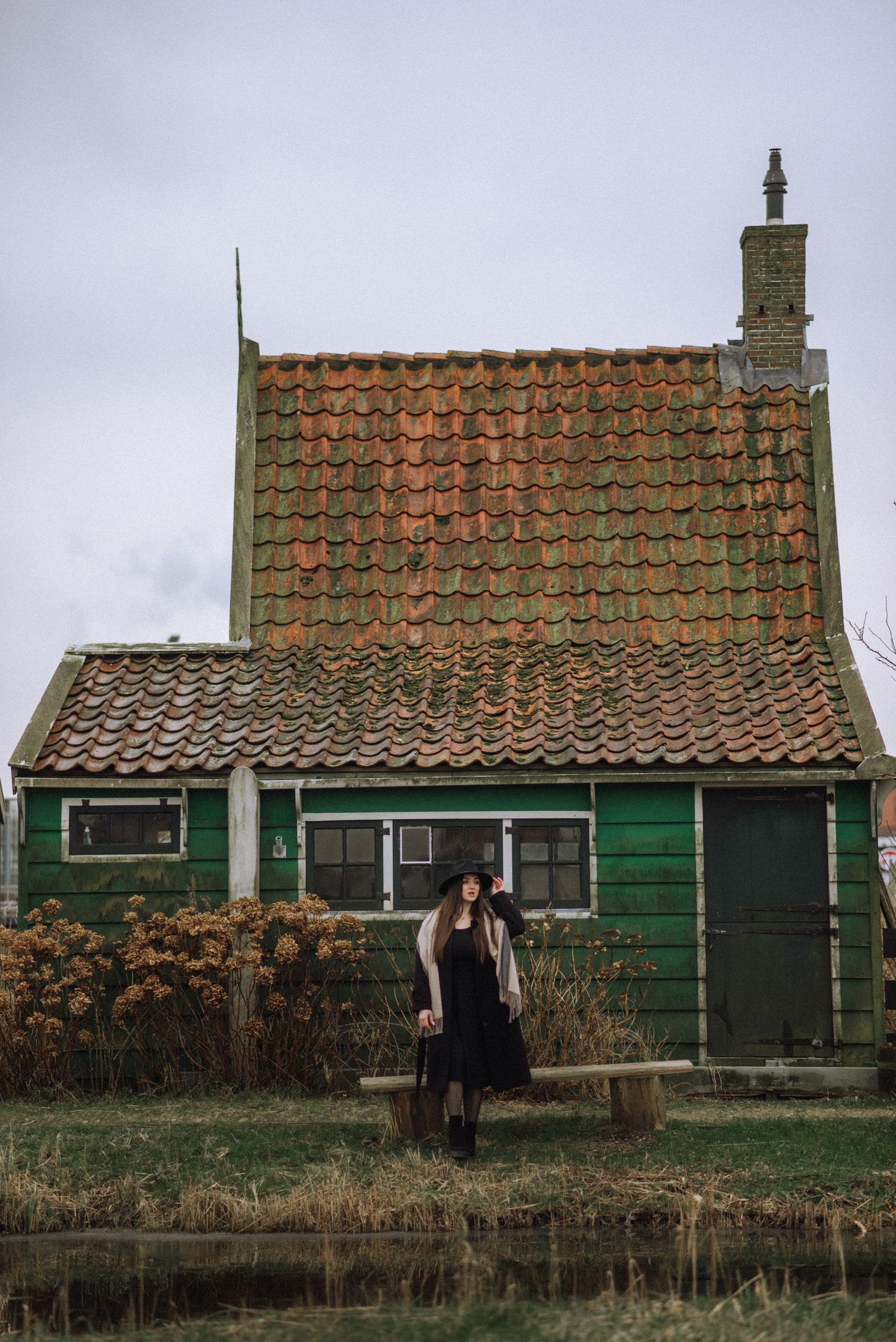 Woman in a black dress and hat stands before a rustic green wooden house with a terracotta tiled roof and brick chimney beside a calm canal.