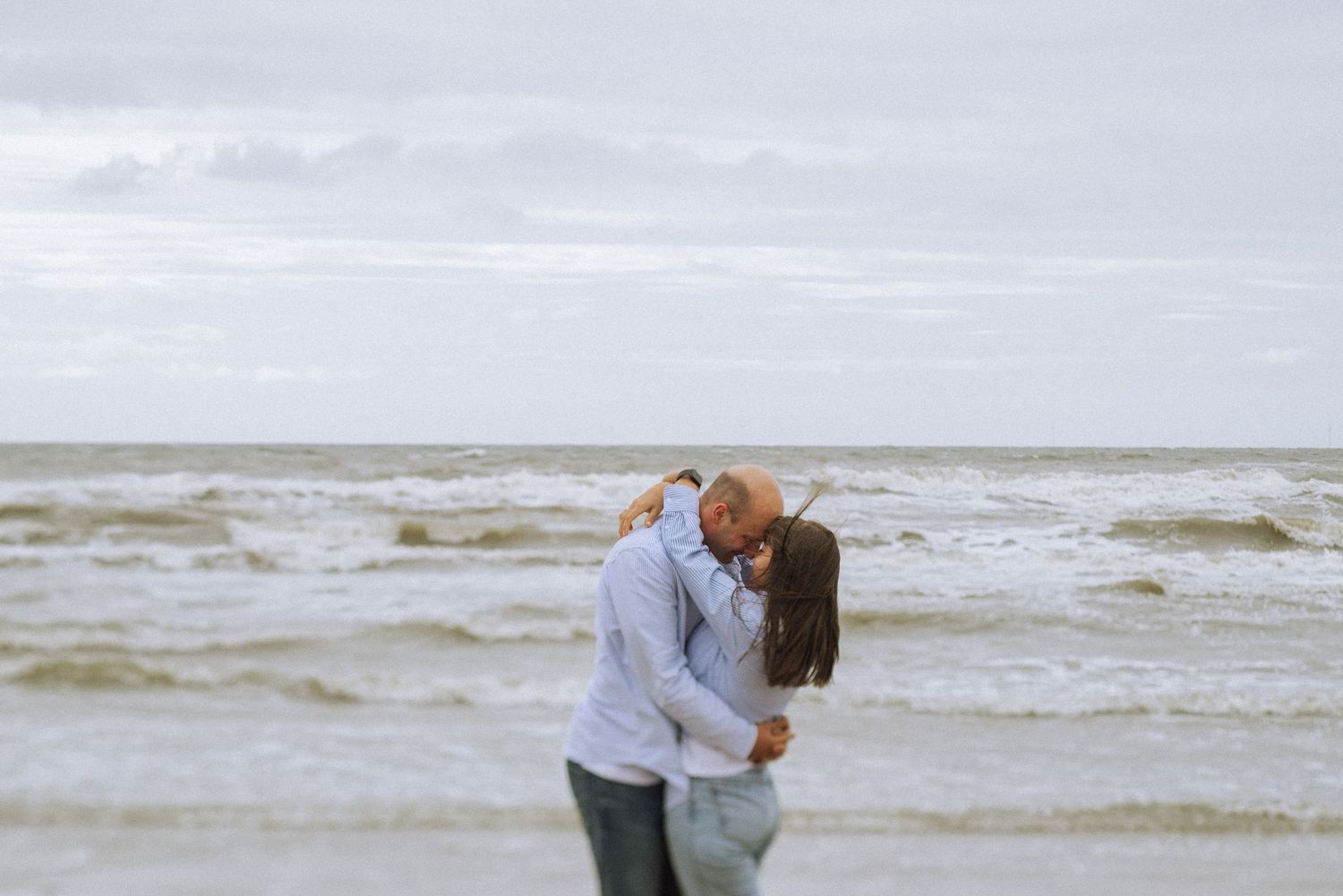 Couple in casual clothes embracing and kissing on a windy beach with rough waves rolling in under a cloudy gray sky.