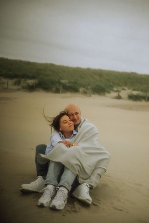 Couple wrapped in a light blanket sitting close together on a windswept sandy beach with grassy dunes under an overcast sky.