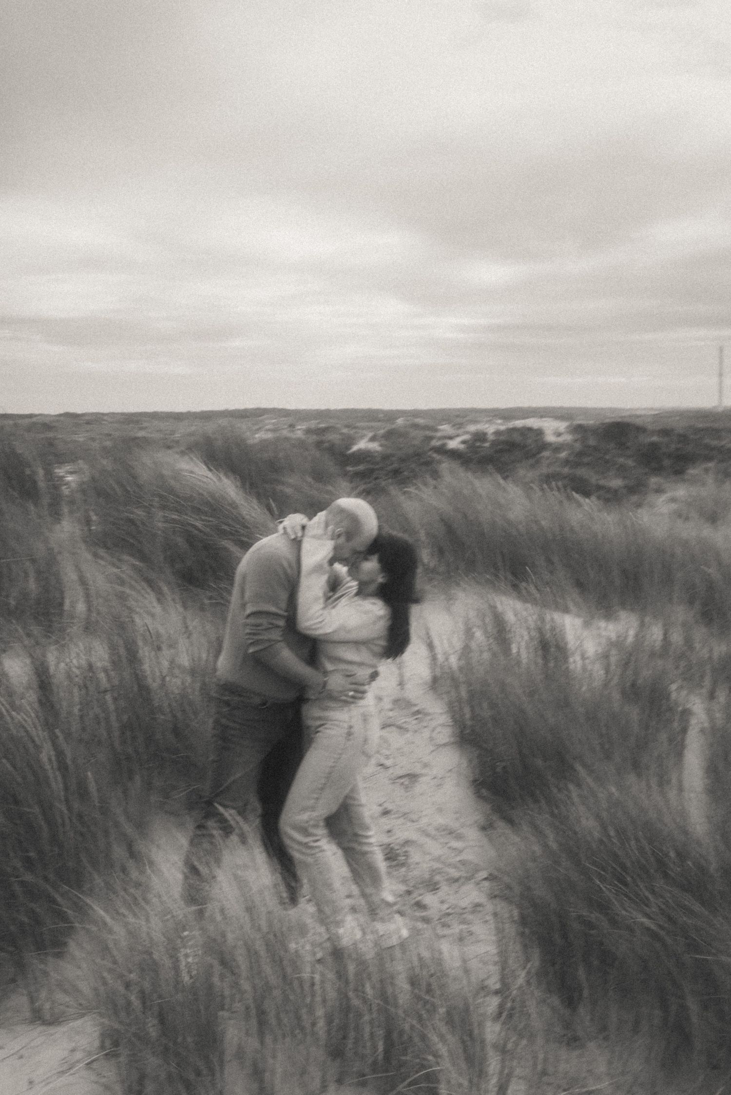 Black and white photo of a couple embracing and kissing on a sandy path through beach dune grass under a cloudy sky.