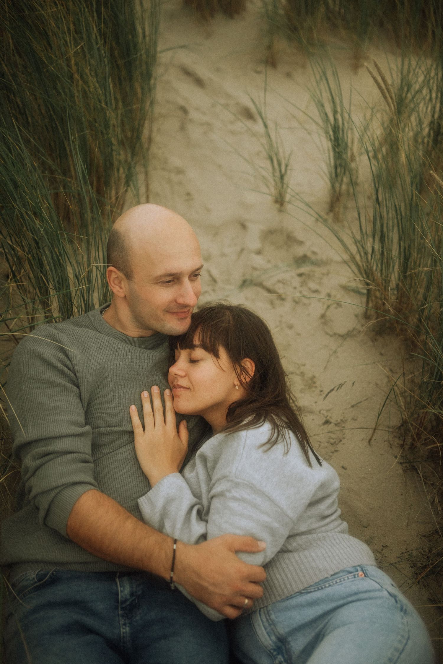 Couple in gray sweaters and jeans embracing and resting together among beach dune grass in a tender, romantic outdoor portrait.