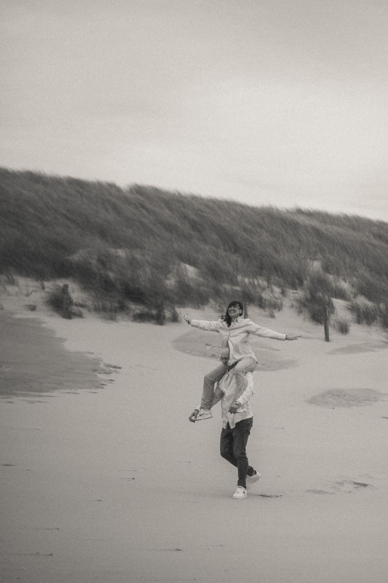 Black and white photo of a person riding on another's shoulders with arms outstretched on a windy sandy beach beside grassy dunes.