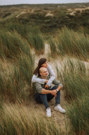 Couple sitting close together on a sandy dune surrounded by tall beach grass, with the woman hugging the man from behind.
