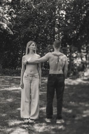 Black and white photo of a barefoot woman in wide-leg jeans and a shirtless man with a back tattoo standing in a sunlit forest clearing.