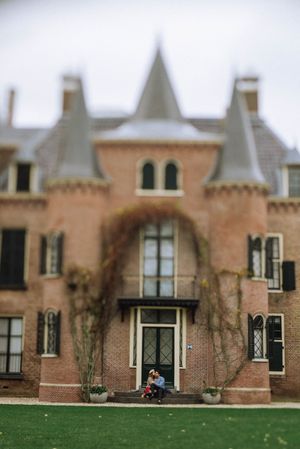 Couple sitting on the front steps of a historic red brick mansion with pointed turrets and ivy-covered walls, set against a green lawn.