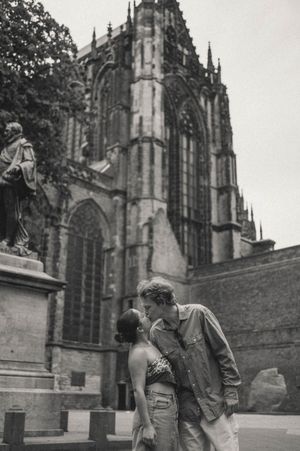 Black and white photo of a couple kissing in front of a gothic cathedral with tall spires and a stone statue on a pedestal.